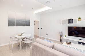 A modern living room with a sofa, dining table, chairs, a flat-screen TV, and artwork on the wall.