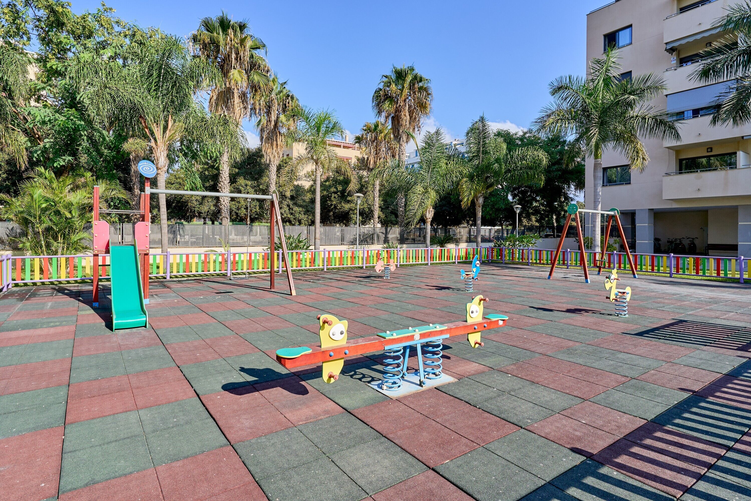 Ein Spielplatz mit Wippen und einer Rutsche, umgeben von Palmen und einem Gebäude.
