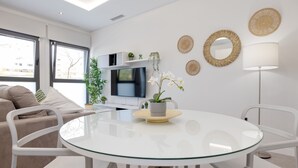 Un salón moderno con una mesa de vidrio, sillas blancas, un sofá, un televisor y decoración mural.