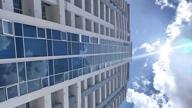 Ein modernes Gebäude mit einer reflektierenden Glasfassade und symmetrischer Gestaltung, ausgestattet mit mehreren Balkonen und blauem Himmel mit Wolken.