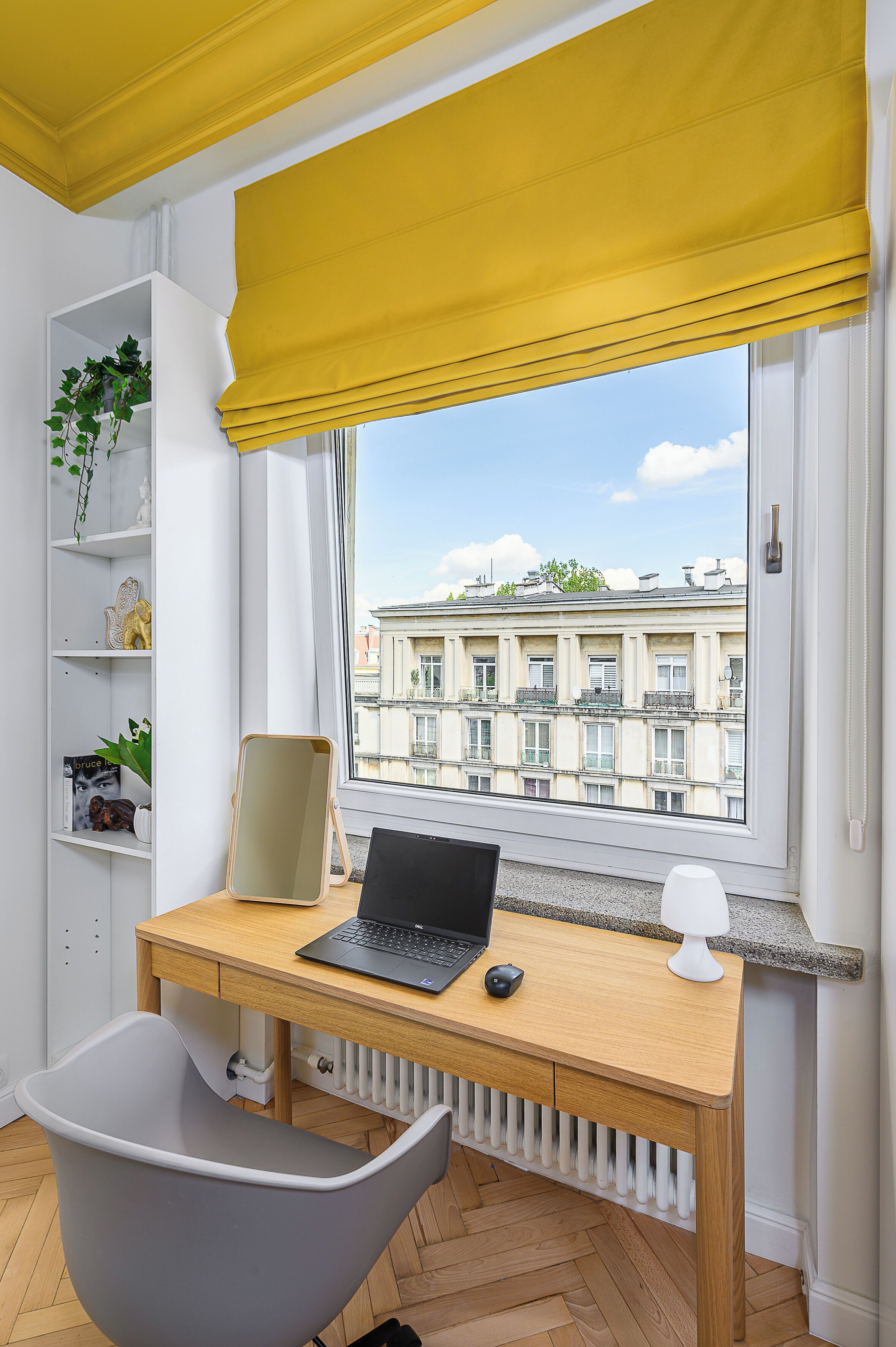 A modern room with a wooden desk, a laptop, and a chair. A window with yellow blinds offers a view of buildings.