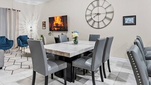 A dining area with a marble table, grey upholstered chairs, a large wall clock, and a TV displaying a fire.