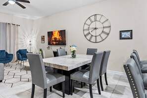 Un comedor con una mesa de mármol, sillas tapizadas en gris, un gran reloj de pared y un televisor mostrando un fuego.