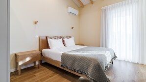 A bedroom with a wooden bed, a bedside table, a nightstand, a wall-mounted air conditioner, and a window with sheer curtains.