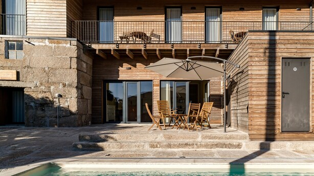 Un espace piscine doté d’une terrasse en bois, de sièges extérieurs et d’un bâtiment aux murs en pierre et aux balcons.