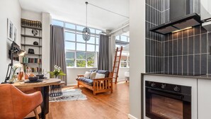 Uma cozinha moderna com forno embutido, área de refeições com mesa e cadeiras de madeira, sofá e estante com livros e objetos decorativos.