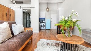 Uma sala de estar moderna com um sofá de couro marrom, uma mesa de centro de madeira com um vaso de flores, e um tapete de couro de vaca branco. Uma cozinha com uma máquina de lavar e uma geladeira azul é visível ao fundo.