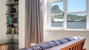 Quarto com um banco de madeira, estante e janela com vista para uma montanha e um corpo d'água.