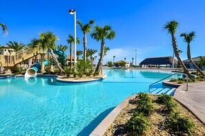 Una gran piscina al aire libre con tobogán de agua, palmeras y un edificio con techo de paja al fondo.