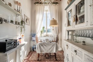 A narrow hallway with a dining table set for two, flanked by white cabinets with glass doors, a chandelier, and a window with sheer curtains.