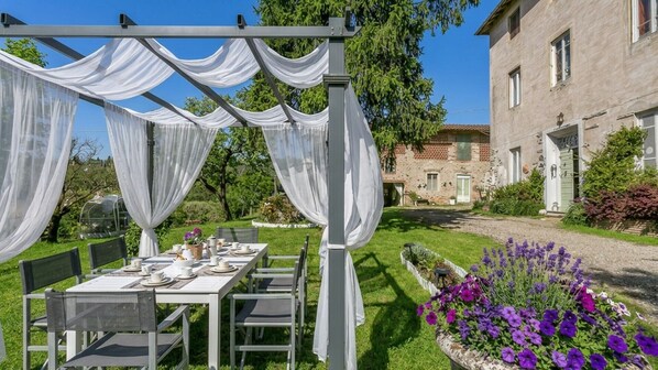 An outdoor dining area with a table set for four, surrounded by white drapes, a stone building in the background, and a garden with purple flowers.