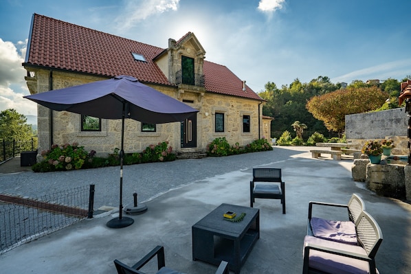 A stone building with a red-tiled roof, outdoor seating with chairs and tables, and a large umbrella.
