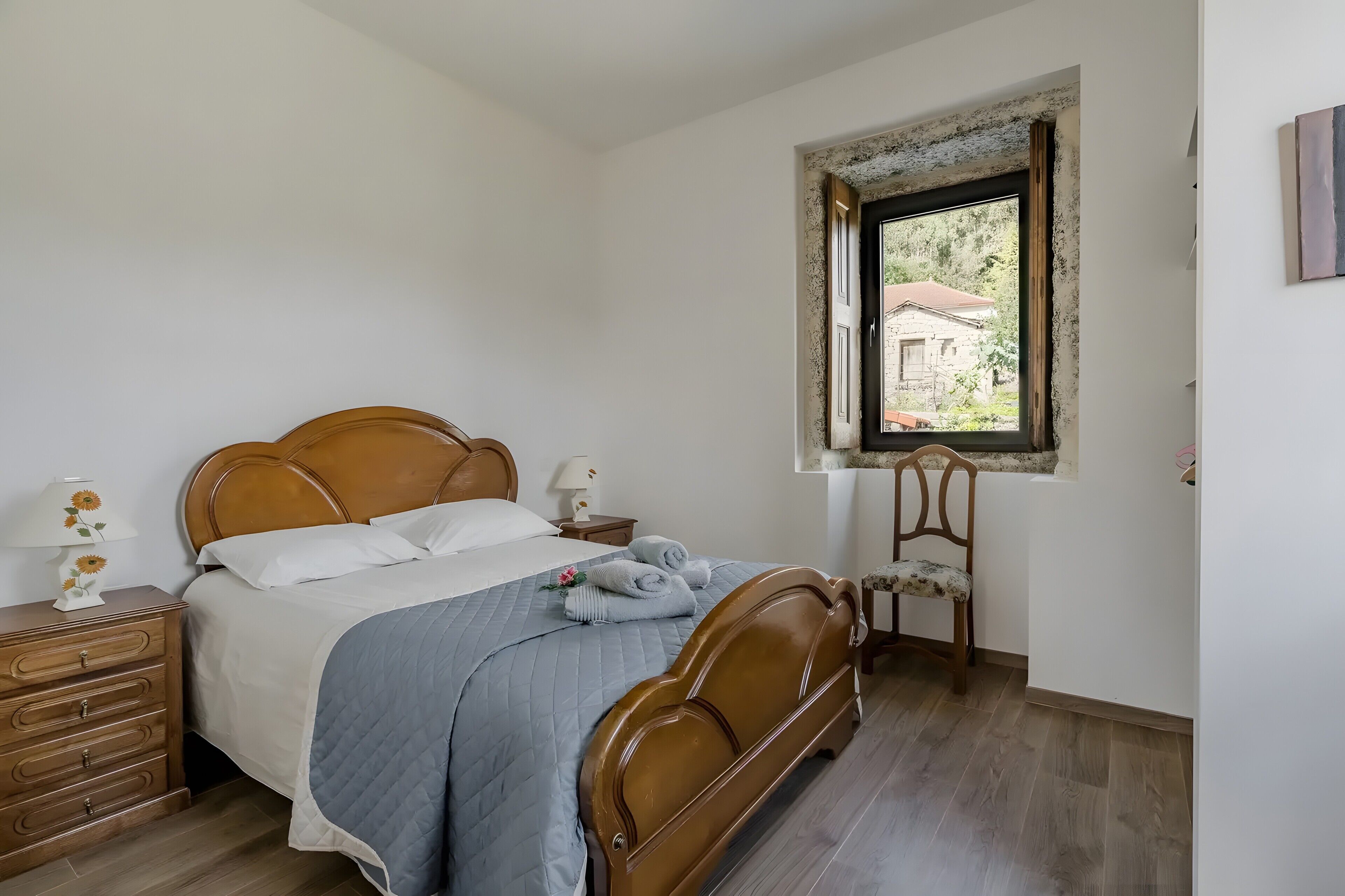 Een slaapkamer met een houten bed, een nachtkastje, een stoel en een raam met uitzicht op groen.