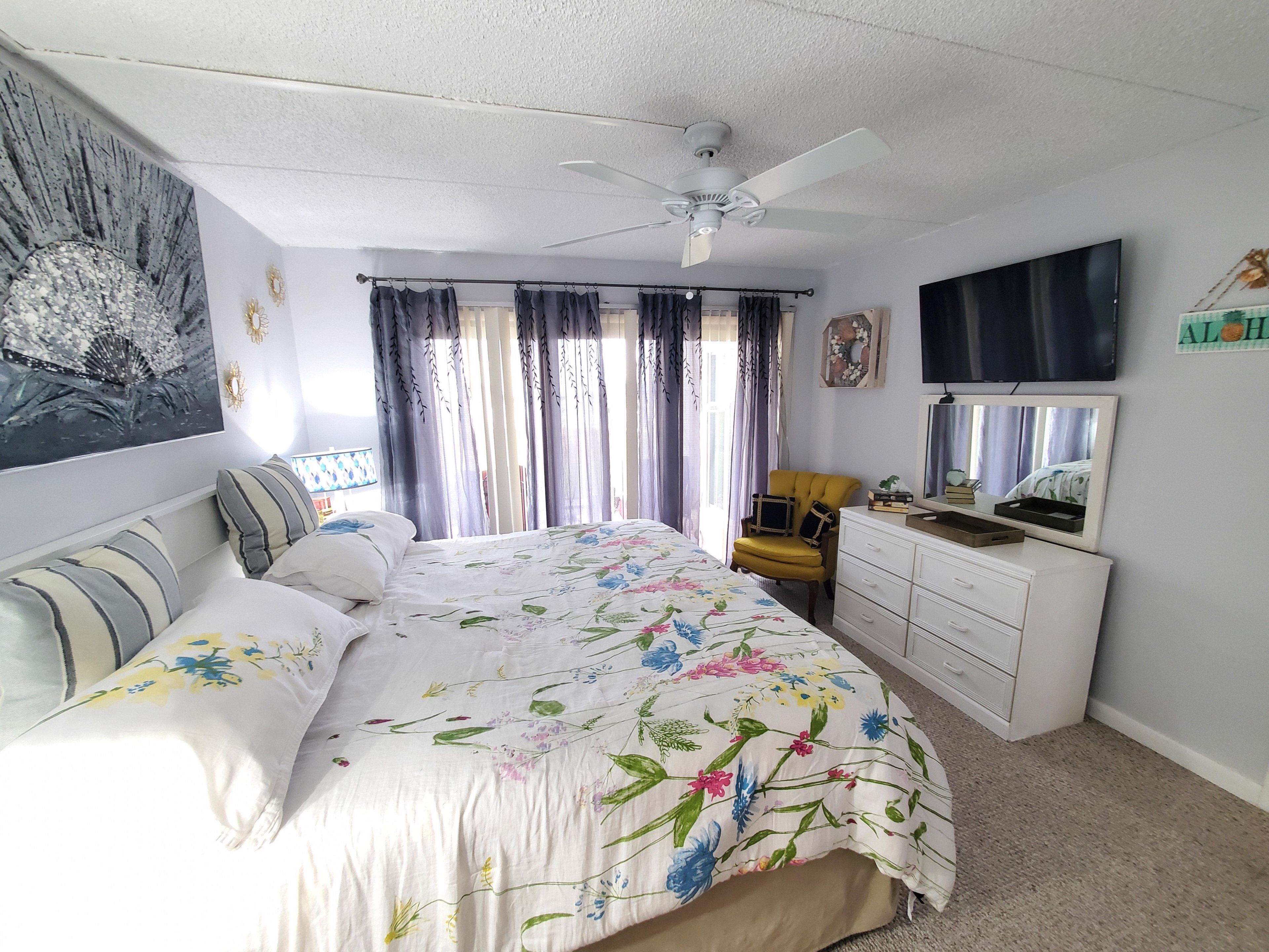 A bedroom with a bed, a chair, a dresser, a ceiling fan, and a TV.