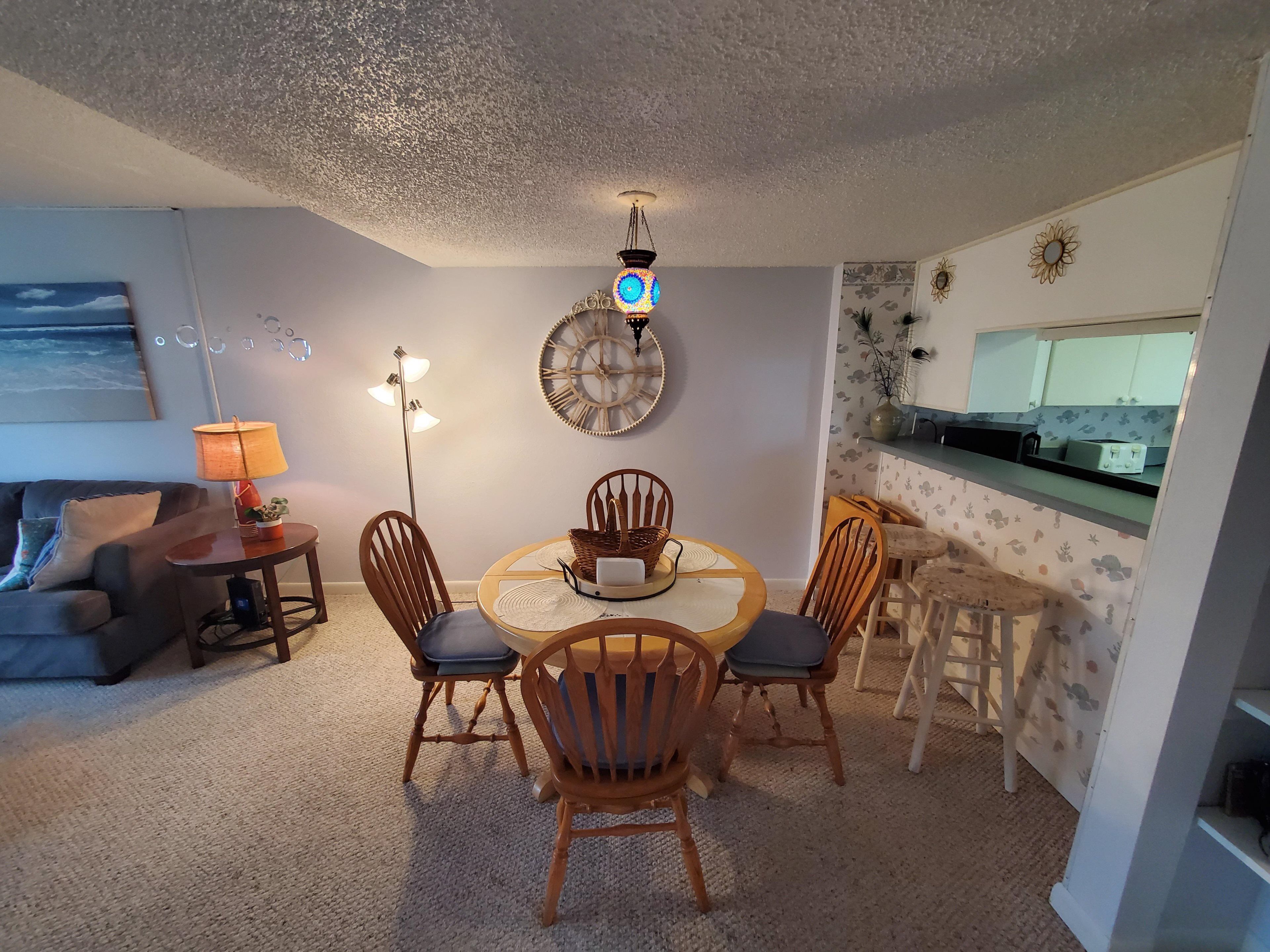 Een gezellige woonkamer met een eethoek, met een ronde tafel, houten stoelen, een vloerlamp en een wandklok.