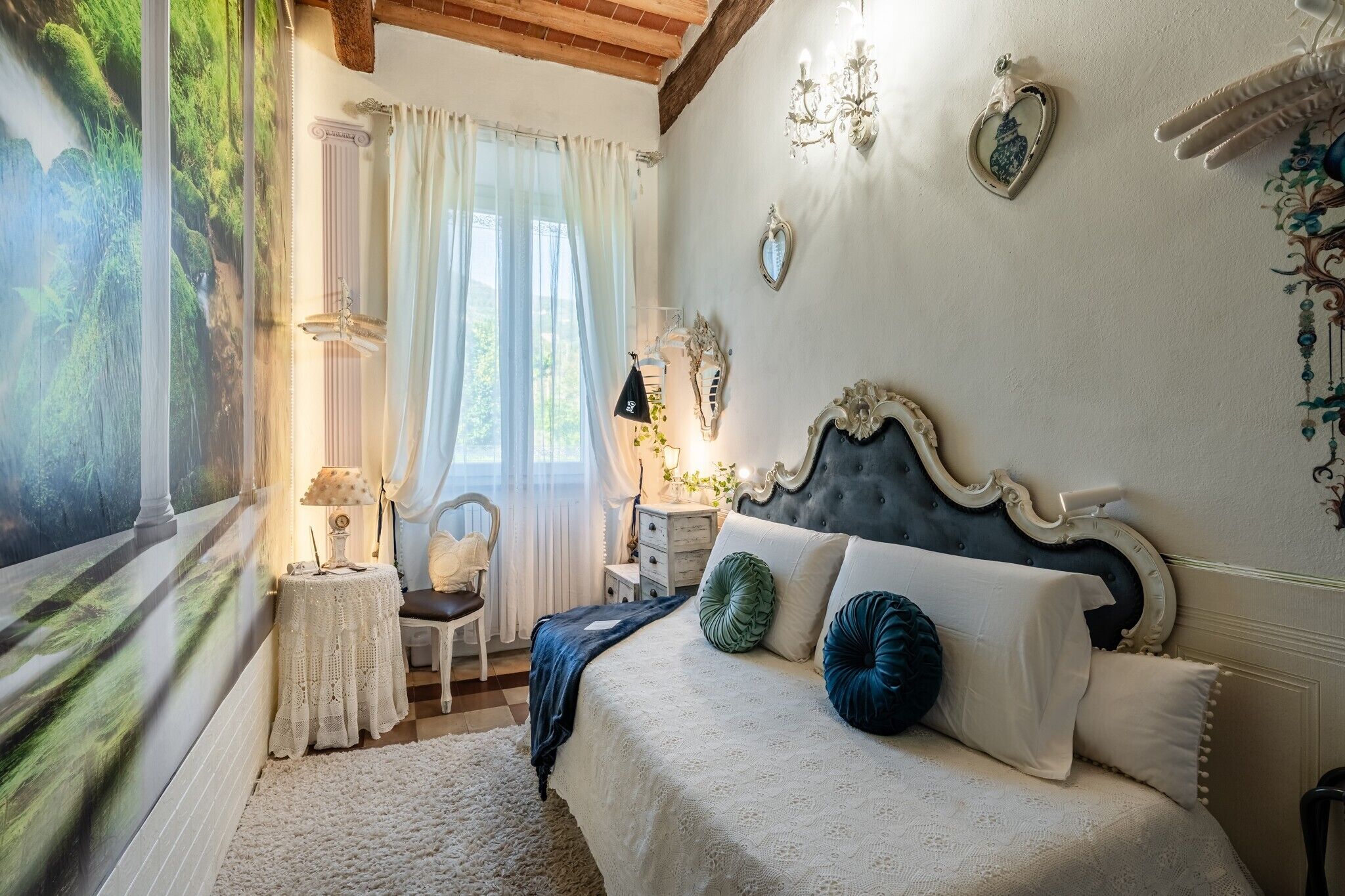 Une chambre à coucher avec un grand lit, une table de chevet, une chaise, une fenêtre avec des rideaux et un mur agrémenté d’éléments décoratifs.