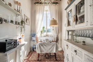 A narrow room with a dining table, chairs, a chandelier, and a window with curtains.