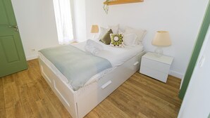 Une chambre à coucher avec un lit, des tables de chevet, une commode, une étagère avec une plante et une porte.