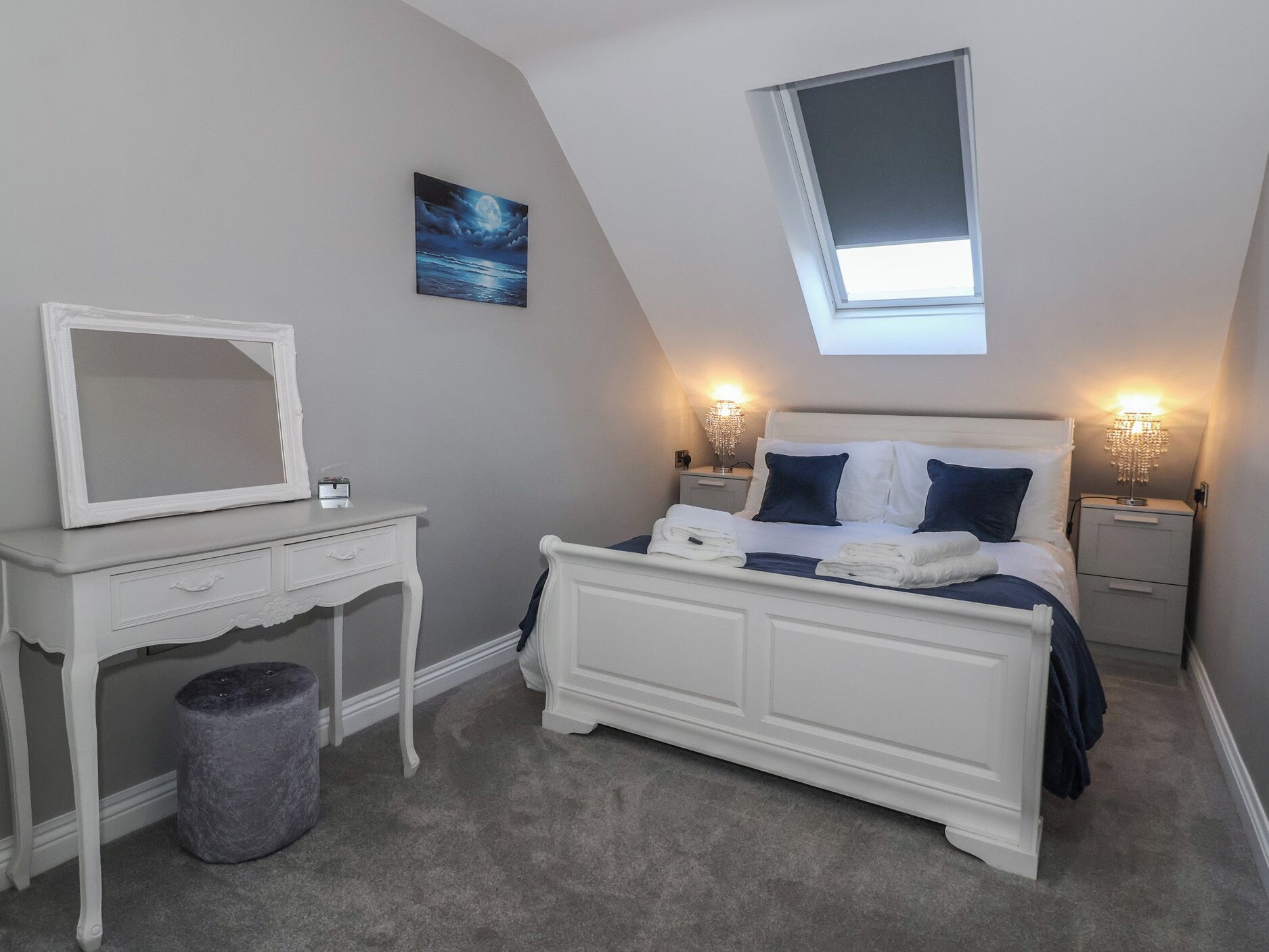 Ein Schlafzimmer mit einem Bett, Nachttischen, einem Spiegel, einem kleinen Hocker und einem Bild an der Wand.