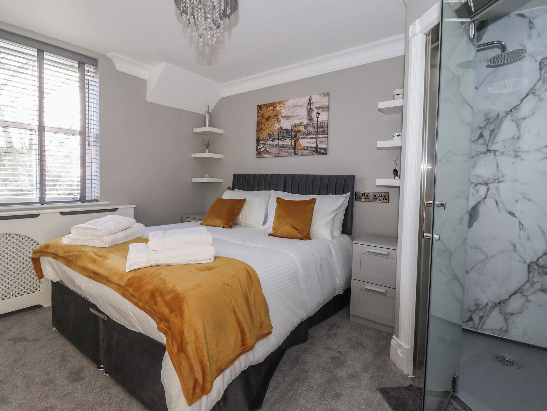 A modern bedroom with a bed, bedside tables, a nightstand, a glass shower enclosure, and a window with blinds.