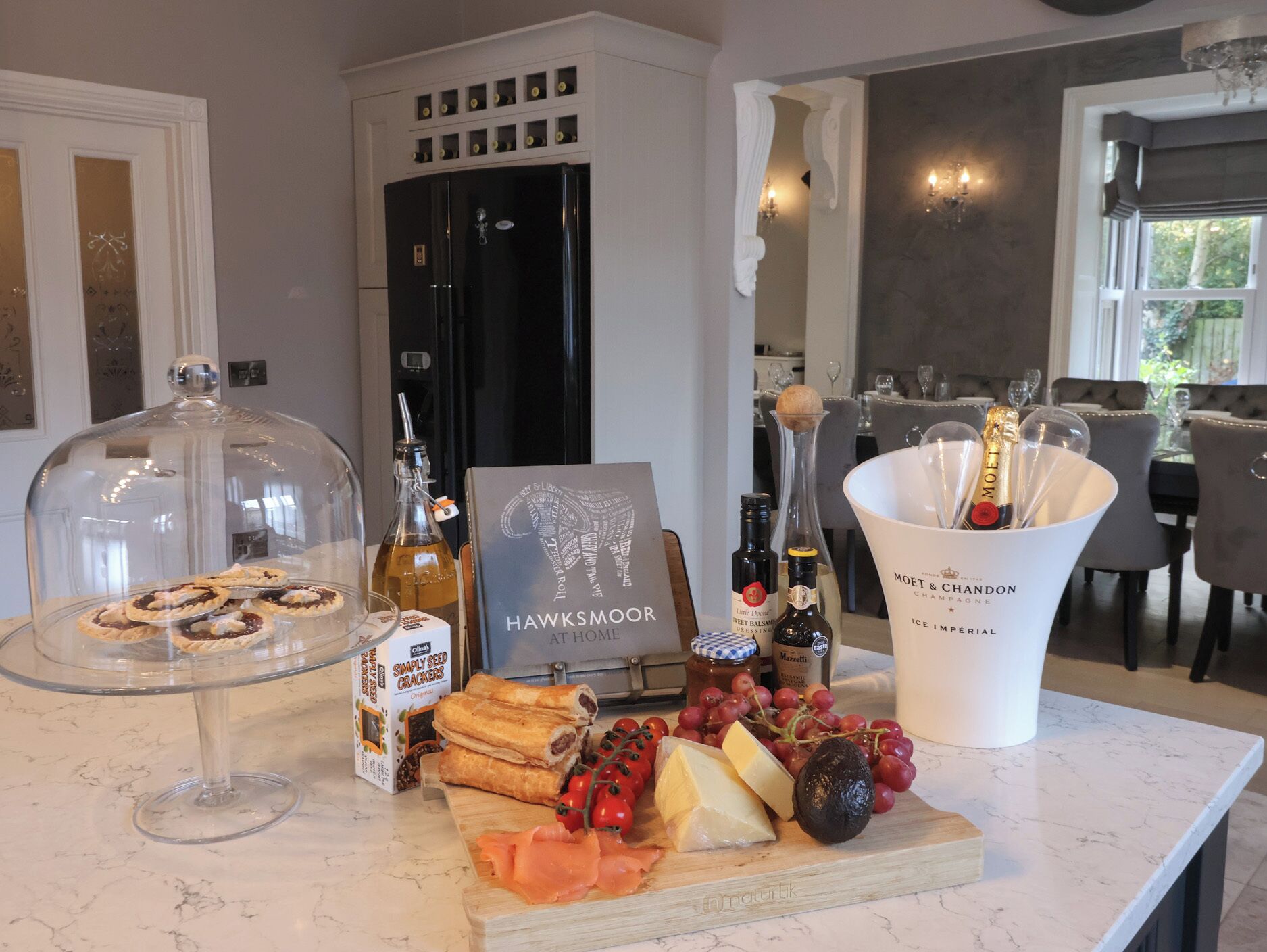 A well-lit kitchen with a marble countertop displaying a variety of food items, including crackers, cheese, and grapes. A black refrigerator and a wine bottle are also visible.
