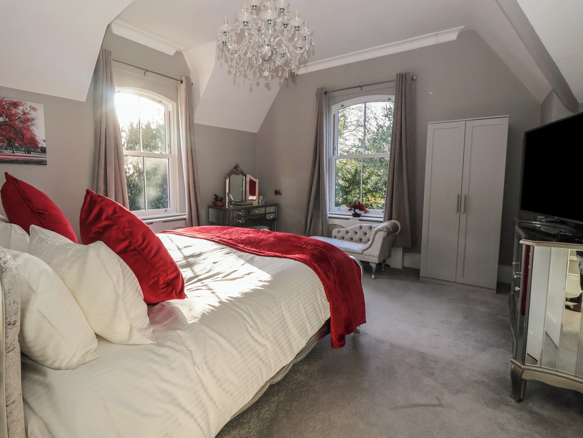 A bedroom with a large bed, two windows, a chandelier, a dresser, and a television.