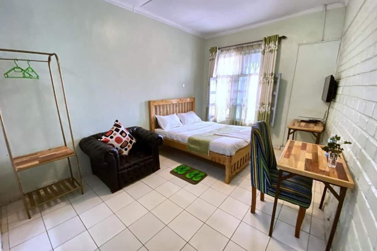A small, clean room with a single bed, a chair, a table, and a hanging clothes rack.