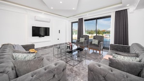 A modern living room with a flat-screen TV, grey sofas, a glass coffee table, and a large window with a view of a pool and mountains. - Villa Grand Yasar 1 With 4 Bedrooms and 4 Bathrooms (Ortaca)