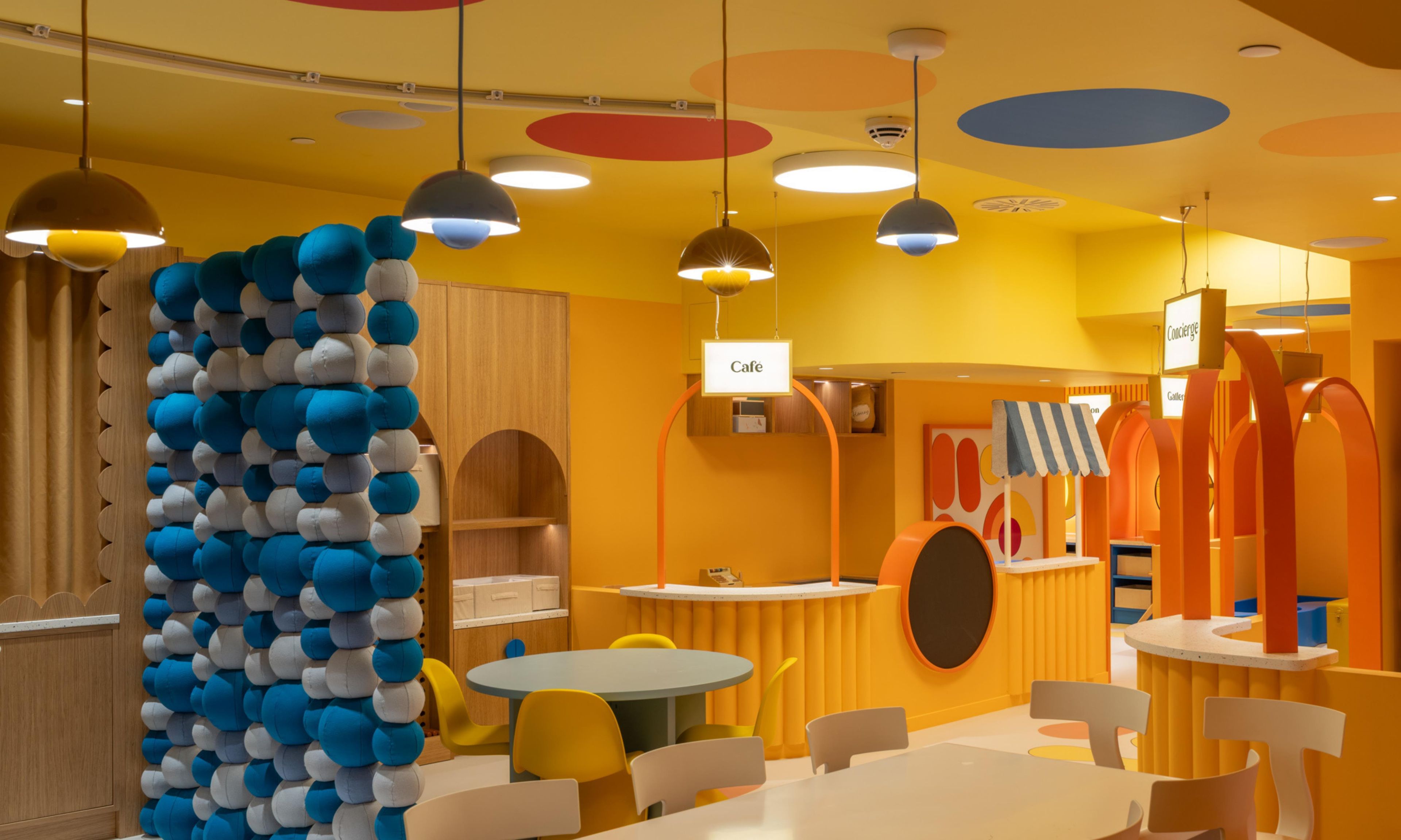 Um espaço interno vibrante, com mesas e cadeiras redondas, uma parede com design esférico azul e branco, e uma placa indicando uma área de café.