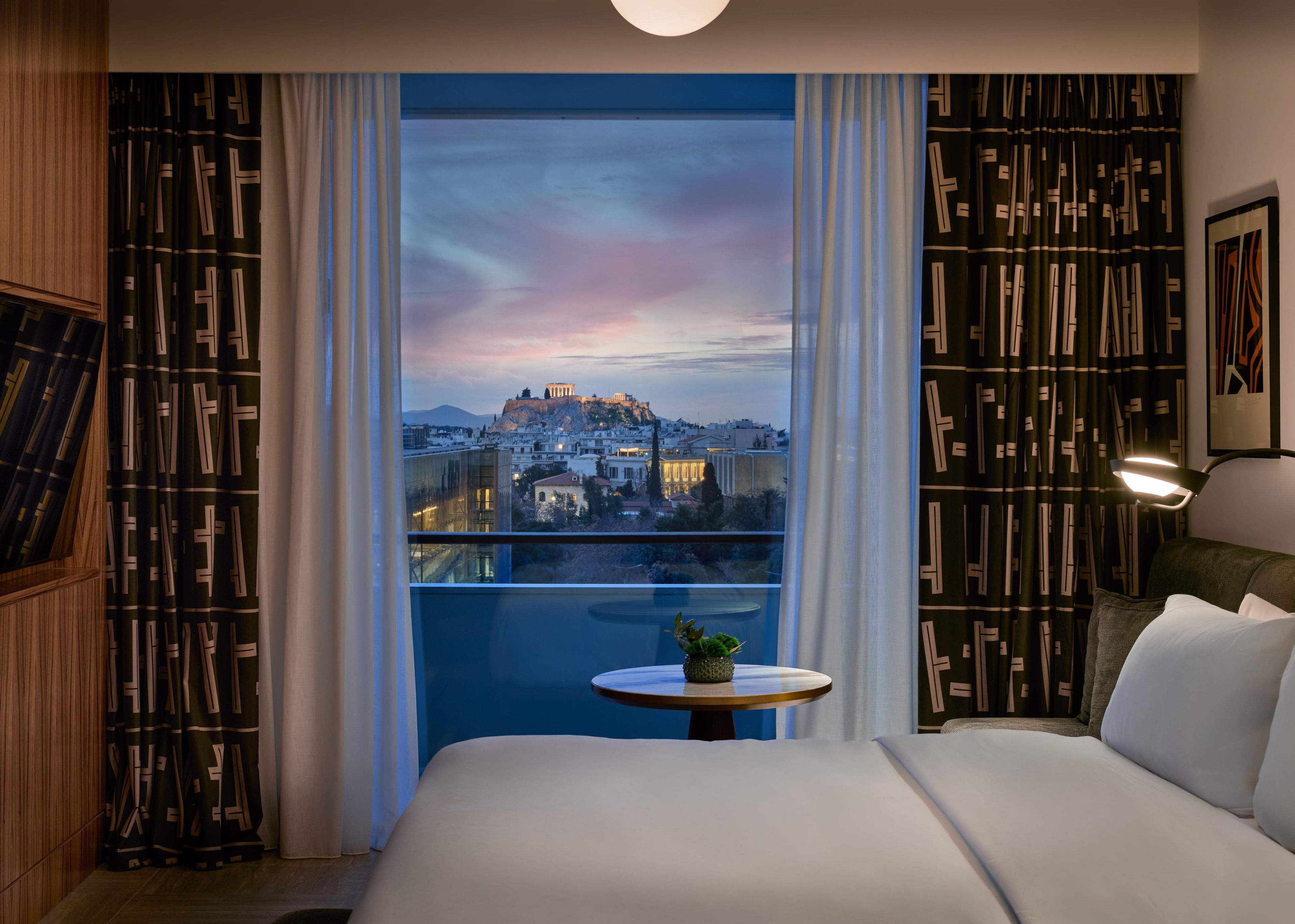 A hotel room with a large bed, a small round table with a plant, and a view of a cityscape with a historic building.