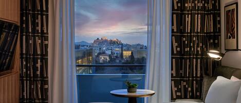 A hotel room with a large bed, a small round table with a plant, and a view of a cityscape with a historic building.