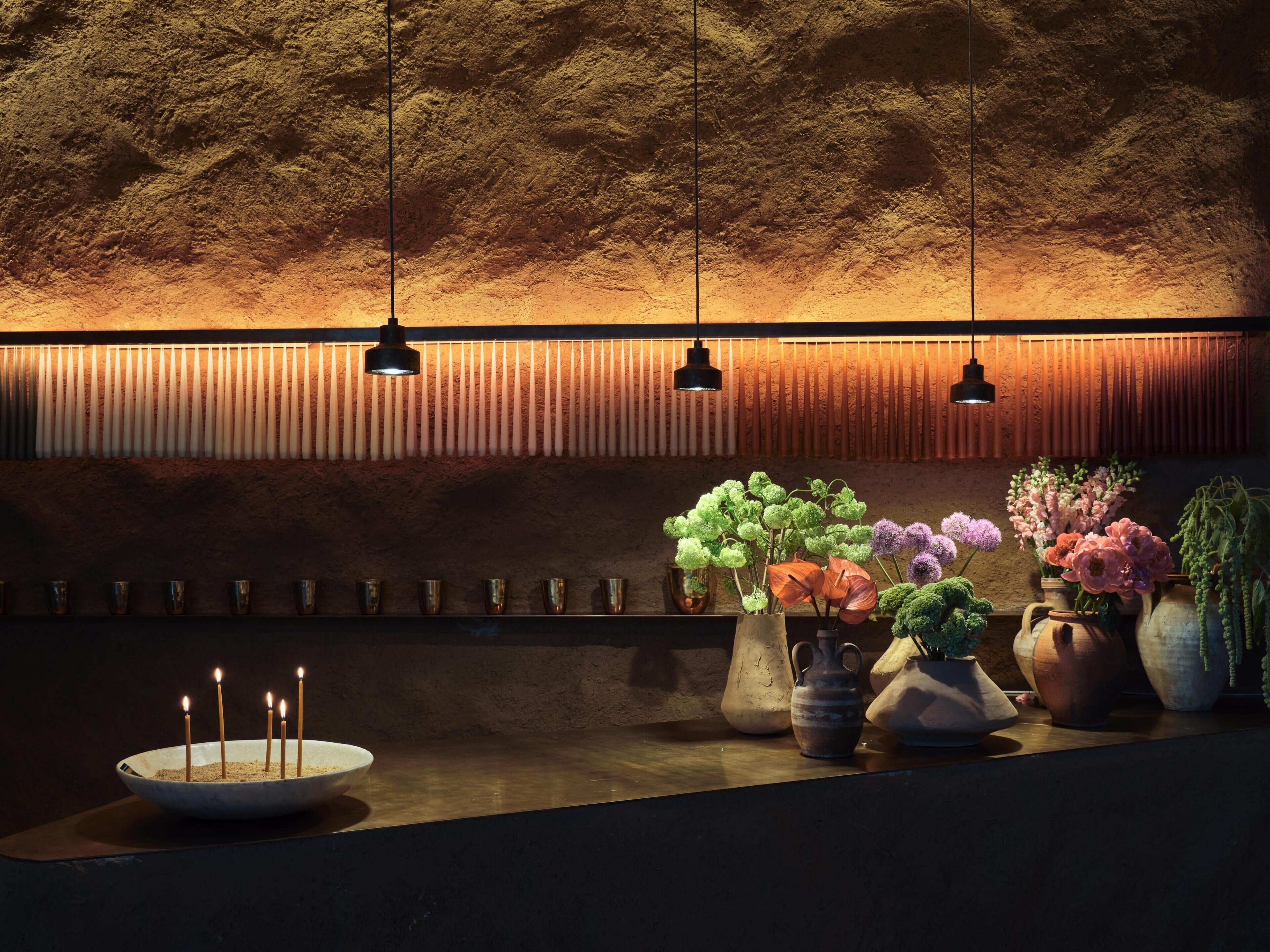 A dimly lit room with a textured wall, a row of candles, and several vases with flowers.