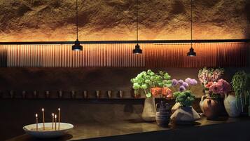 A dimly lit room with a textured wall, a row of candles, and several vases with flowers.