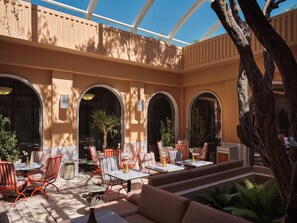 An outdoor seating area with tables and chairs, a large tree, and a covered section with arches. - Conrad Athens The Ilisian (Athens)