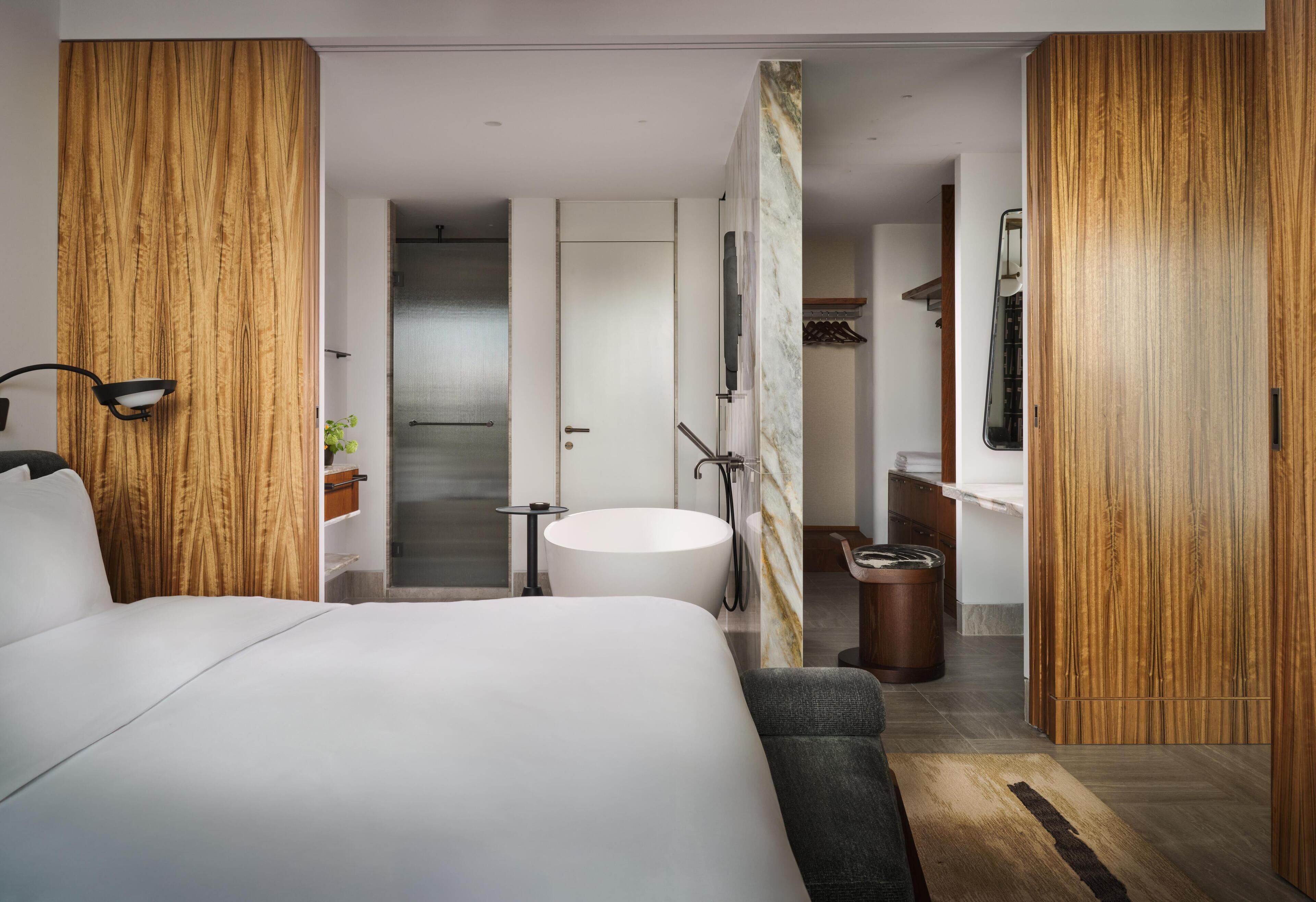 A modern hotel room with a large bed, a round bathtub, a wooden accent wall, and a small seating area.