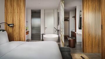 A modern hotel room with a large bed, a round bathtub, a wooden accent wall, and a small seating area.