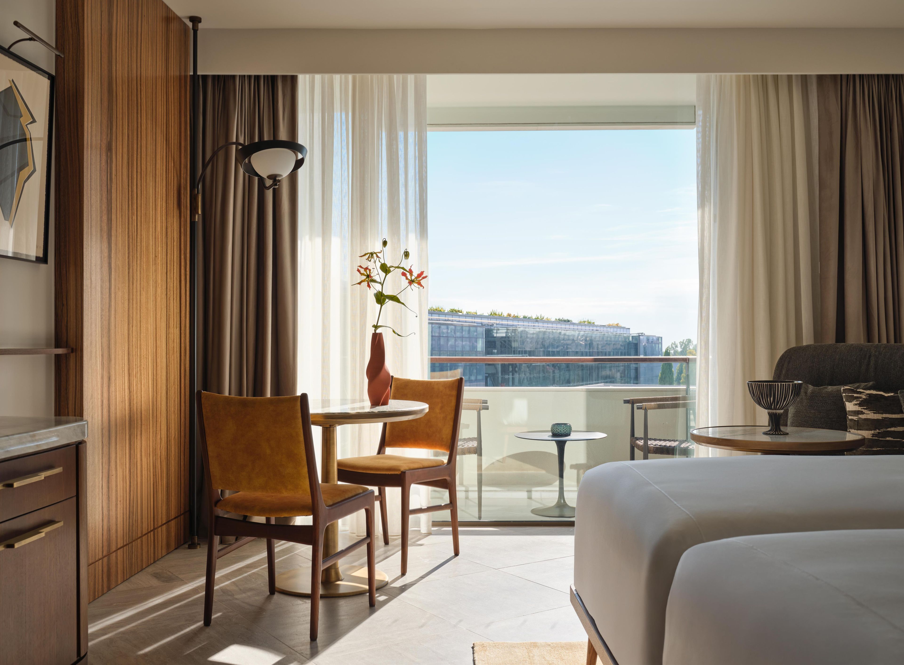 Une chambre d’hôtel avec un lit, un bureau, deux chaises, une petite table, un vase avec des fleurs et une vue sur la ville.
