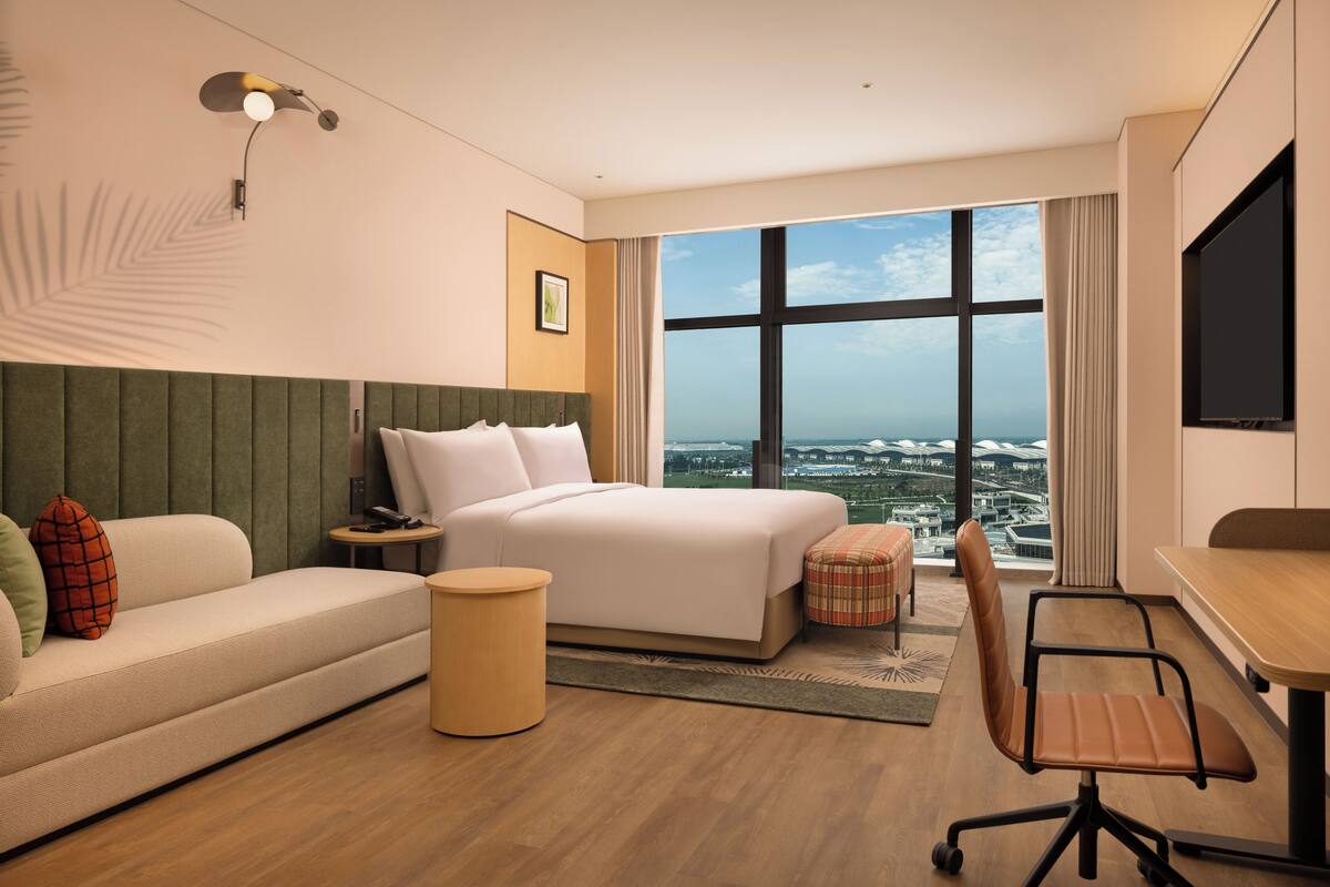 A hotel room with a bed, a sofa, a desk, a chair, a small table, a lamp, a picture on the wall, and a large window with a city view.