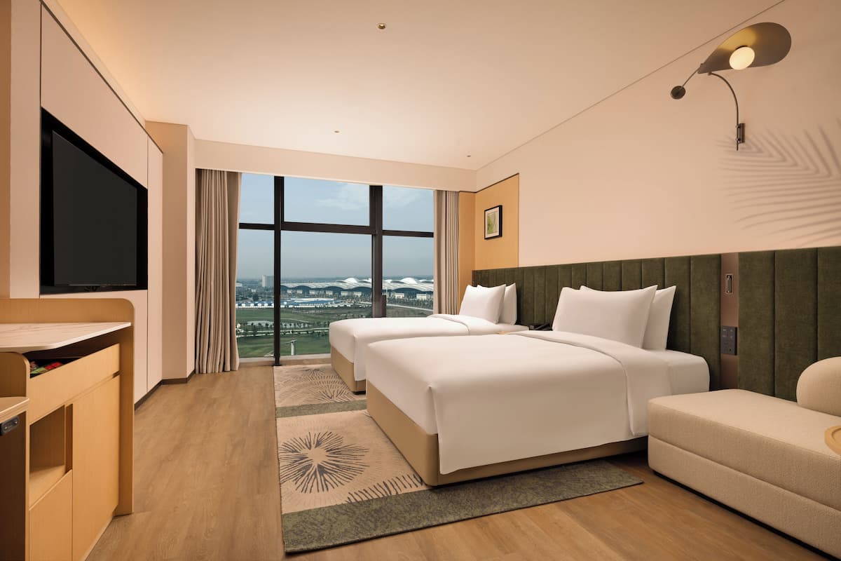A hotel room with a large window, two beds, a TV, a sofa, and a desk.