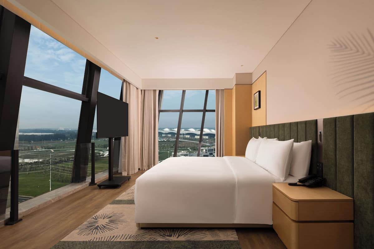 A modern hotel room with a large bed, a nightstand, a TV, and a view of a cityscape through large windows.