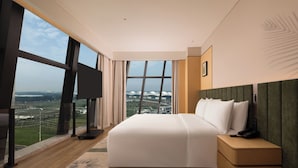 A modern hotel room with a large bed, a nightstand, a TV, and a view of a cityscape through large windows. - Hilton Garden Inn Zhengzhou Airport Aerotropolis (Zhengzhou)