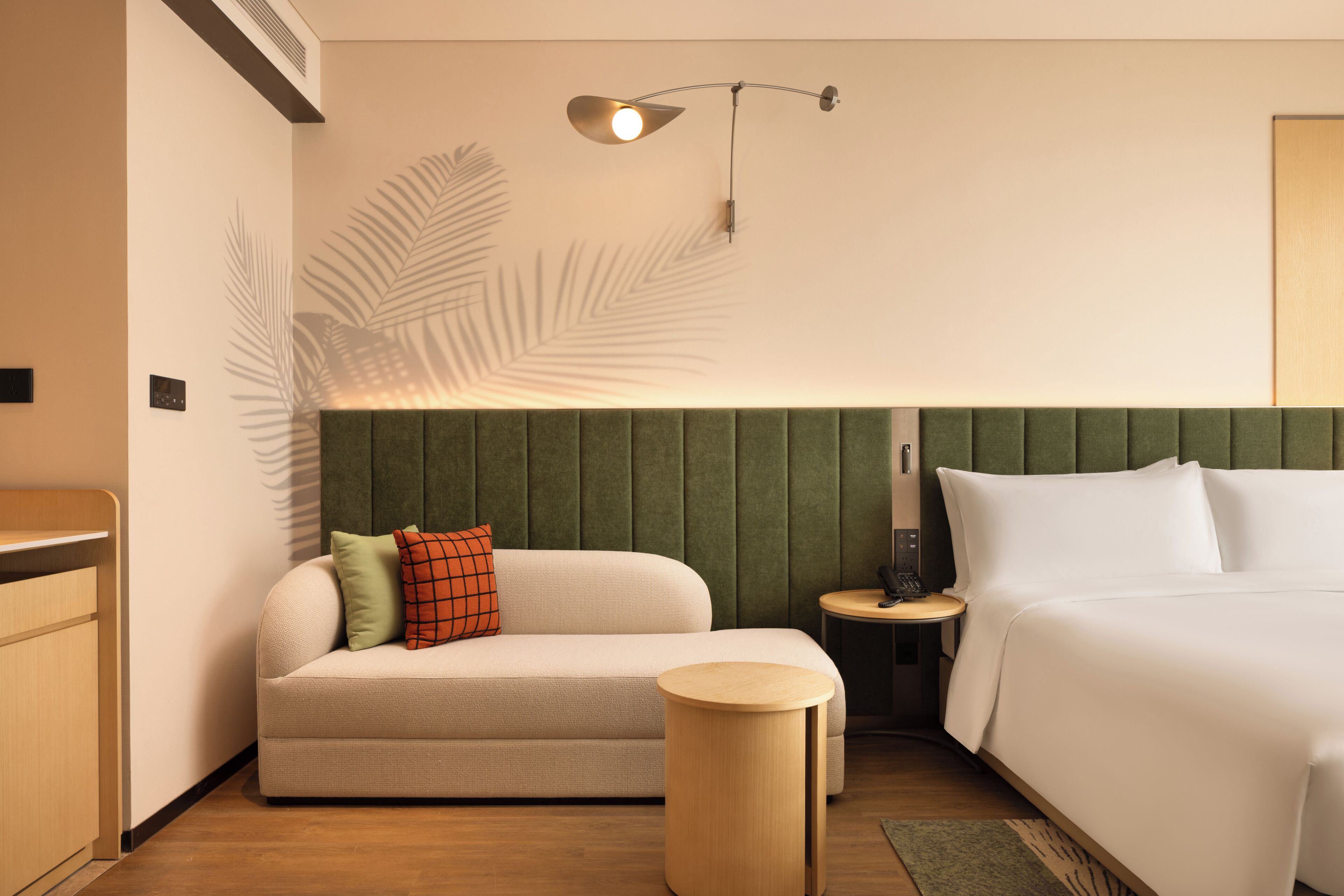 Een hotelkamer met een bed, een bank, een ronde houten bijzettafeltje, een wandlamp en een decoratieve palmbladschaduw aan de muur.