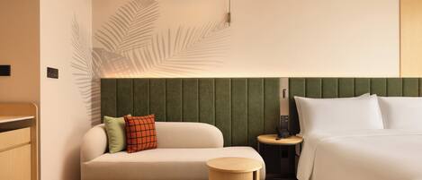Une chambre d’hôtel avec un lit, un canapé, une petite table d’appoint ronde en bois, une lampe fixée au mur et une ombre décorative en forme de feuille de palmier projetée sur le mur.