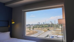 Ein Hotelzimmer mit großem Fenster und Blick auf eine Stadtlandschaft, darunter Straßen, Gebäude und Grünflächen.