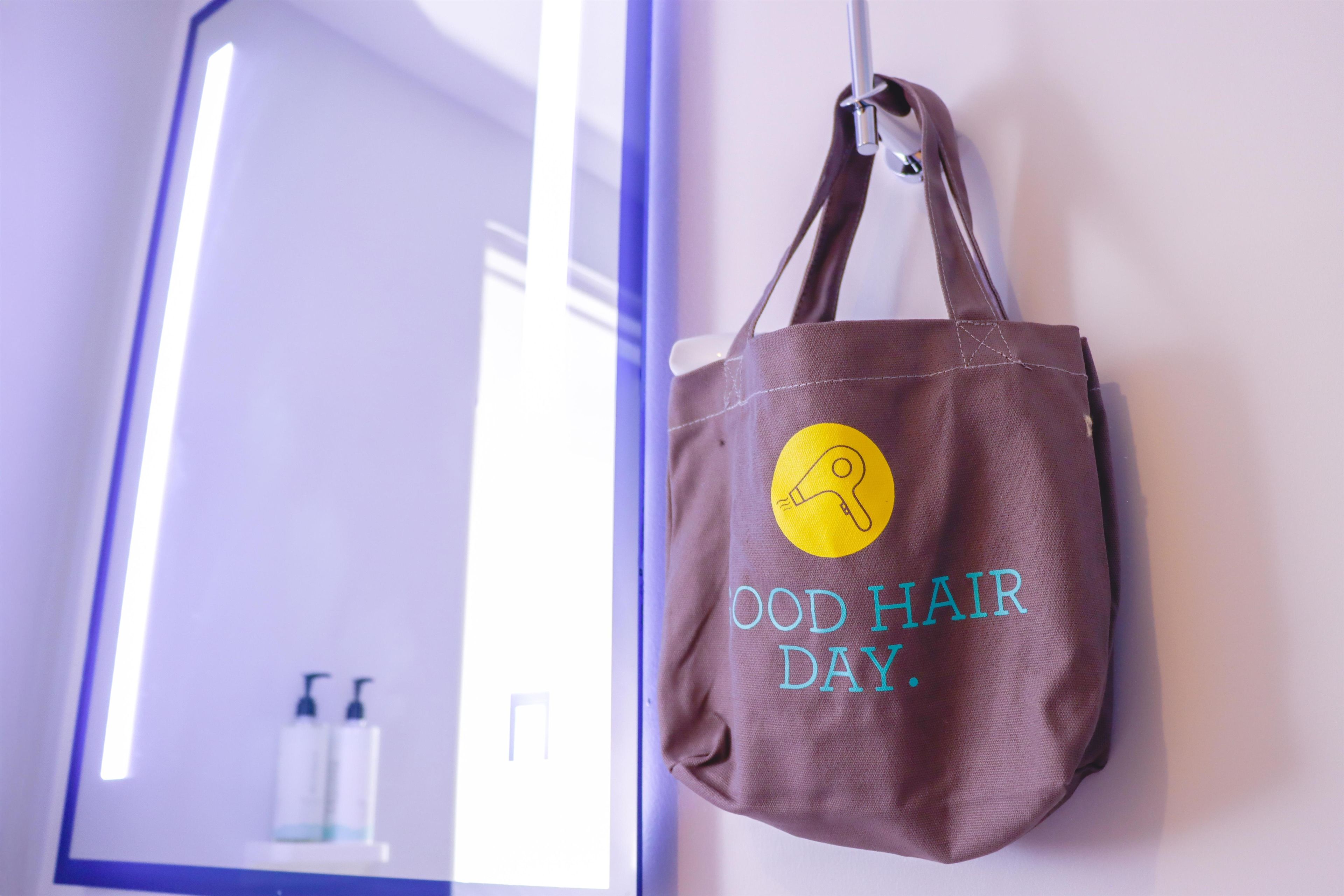 A brown tote bag with a yellow logo and the text 'GOOD HAIR DAY.' hanging on a wall.