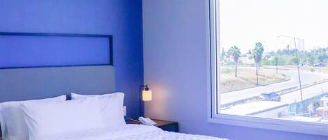 A hotel room with a bed, a nightstand, a lamp, a window with a view of a street and buildings, and a fire alarm on the ceiling.