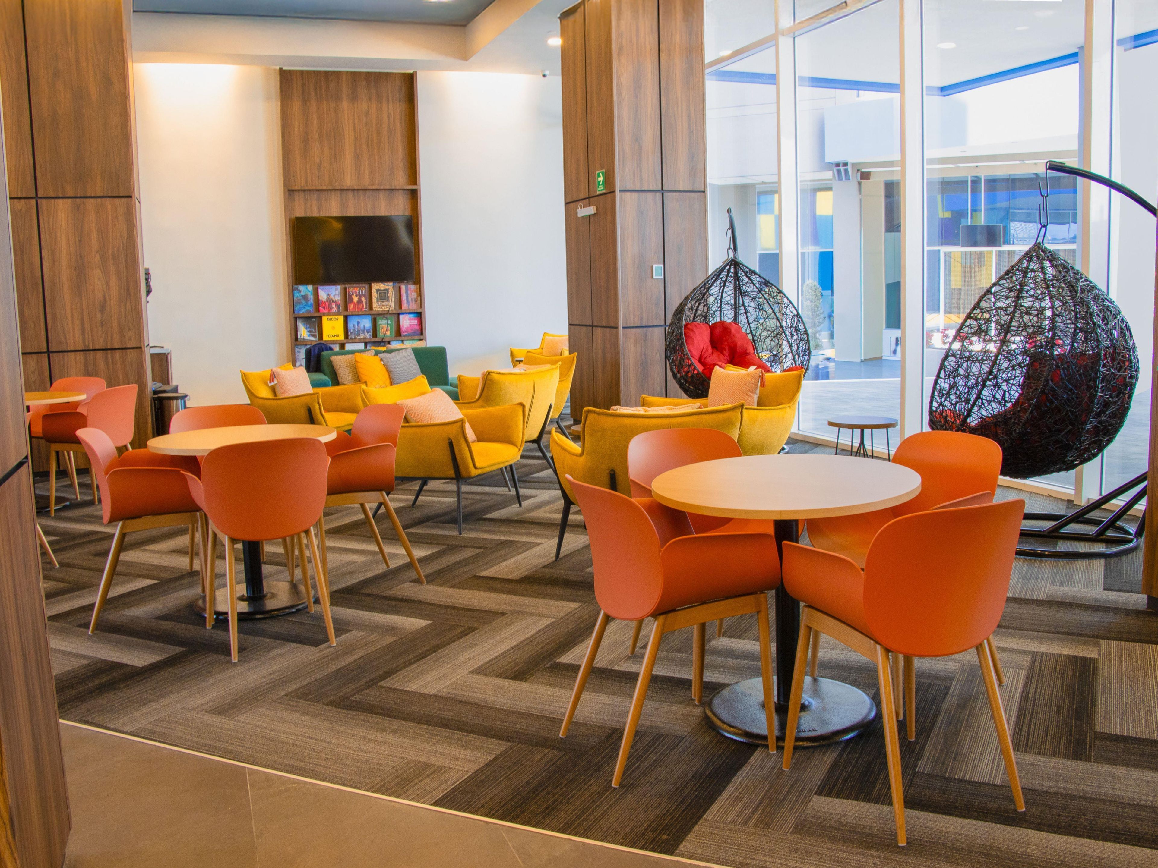 Un espacio lounge moderno con mesas redondas, sillas naranjas, una silla colgante y una estantería llena de libros.