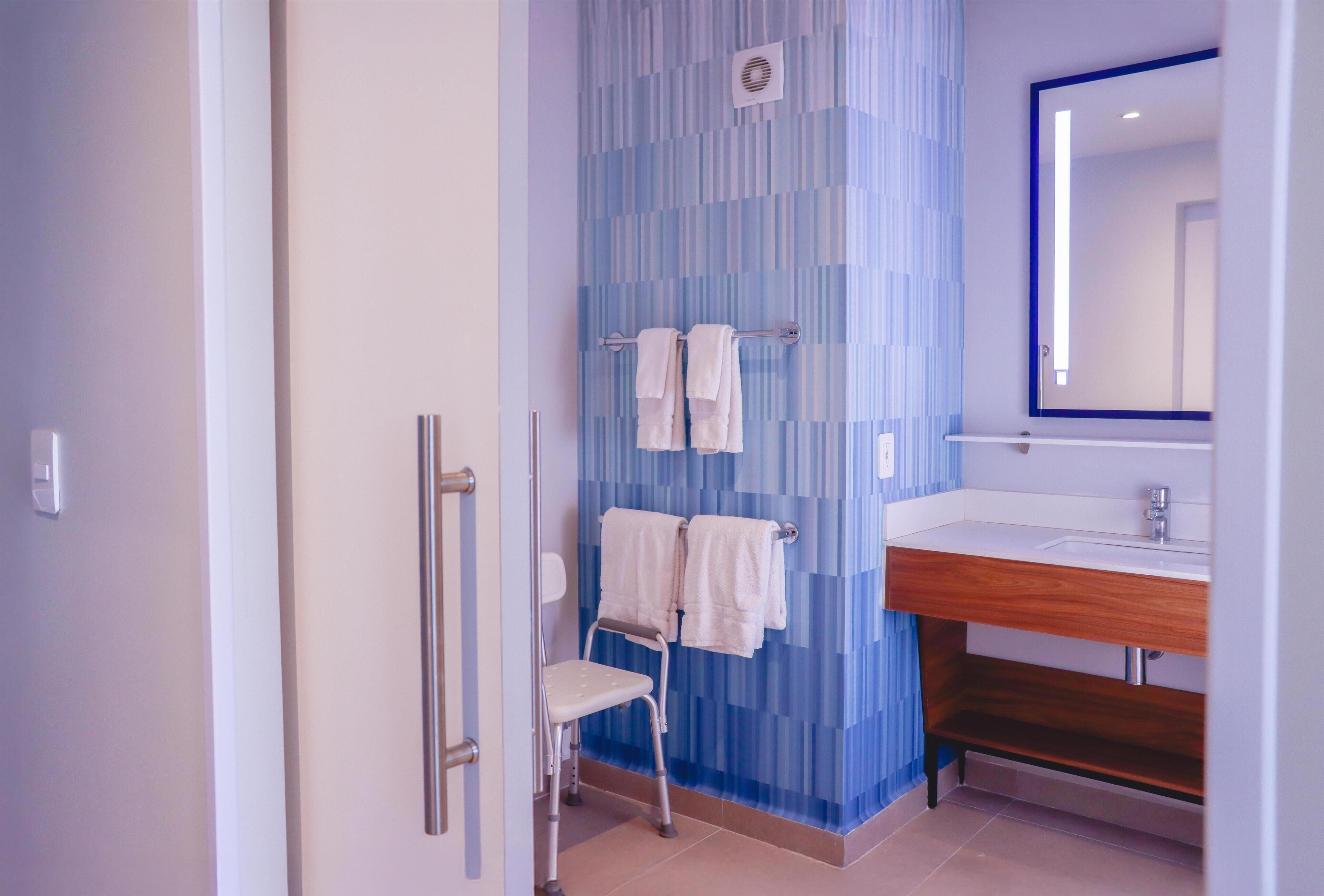 Un baño con pared de azulejos azules, tocador de madera, espejo y una silla accesible para sillas de ruedas.