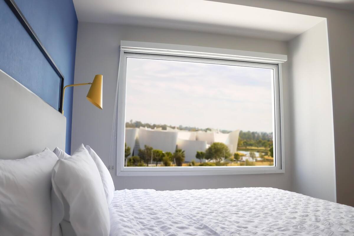 Un dormitorio con un ventanal grande, una cama con ropa blanca y una pared azul.
