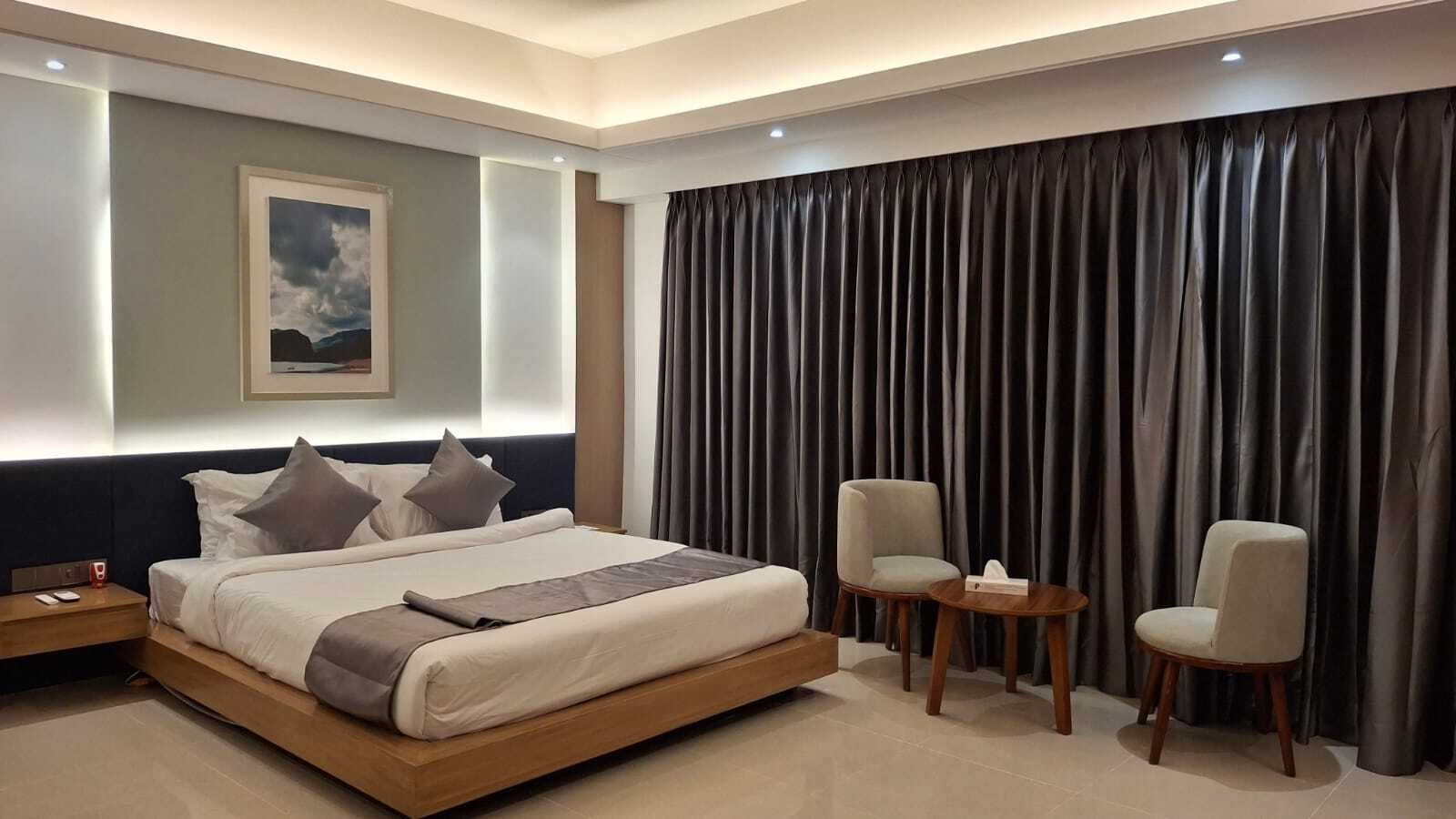 A modern hotel room with a large bed, two armchairs, a small table, and a large window with curtains.